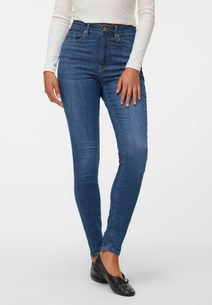 Vrouw draagt blauwe skinny jeans, witte top met lange mouwen en zwarte ballerina's en staat tegen een witte achtergrond.