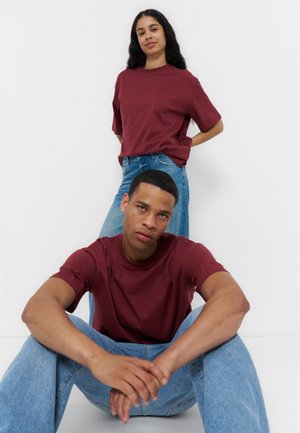 Homme assis par terre les mains jointes, portant une chemise bordeaux et un jean ; femme debout derrière lui les mains dans le dos, même tenue.