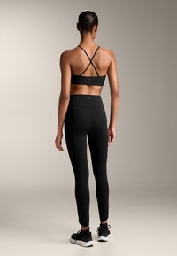 Črn športni modrček s prečnimi naramnicami, visoko pasne črne legice, oboje eleganten in prilegajoč, v kombinaciji s črnimi športnimi čevlji.