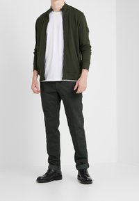 Zelená bomber bunda na zip přes bílé tričko, doplněná tmavými vzorovanými kalhotami a černými botami. Má žebrované manžety a límec.