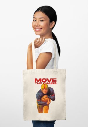 Žena se usmívá, drží bavlněnou tašku na rameno s ilustrací fotbalisty ve vintage stylu a červeným nápisem „POSUŇ ŘETĚZY.“