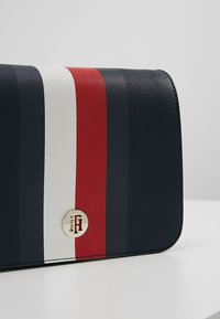 Bolso de mano azul marino con franjas diagonales rojas y blancas. Presenta un acento de hardware con logo dorado en un diseño estructurado. Material texturizado.