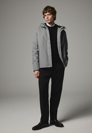 Chaqueta gris con capucha de textura suave, con dos bolsillos frontales y cremallera. Lleva puesta sobre un suéter negro y pantalones a rayas. Zapatos negros.