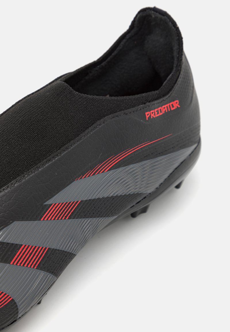 Botín de fútbol negro con un diseño elegante, que presenta un material sintético texturizado, detalles en rojo y franjas grises.