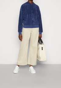 Felpa di velluto blu marina con cappuccio e cordoncino, pantaloni larghi color crema e sneakers bianche. Borsa a tracolla in tela con manici neri tenuta in mano.