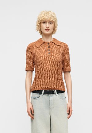 Persona vistiendo una camisa polo de punto color óxido con textura y manga corta, metida dentro de unos jeans de lavado claro con un cinturón negro, de pie sobre un fondo neutro.