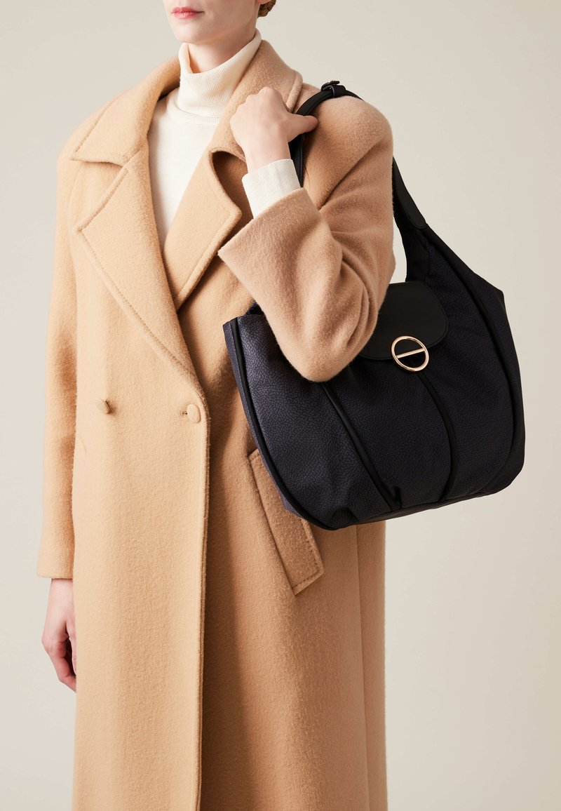 Navy textured shoulder bag with a rounded shape; features a black flap and gold circular hardware. Model wears a camel overcoat.