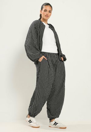 Femme debout portant une veste et un pantalon larges à motif quadrillé noir et blanc, une chemise blanche et des baskets blanches avec des bandes noires.