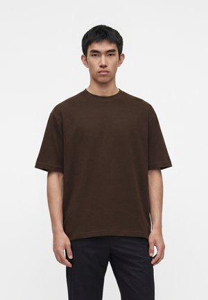 Jeune homme aux cheveux noirs courts, portant un T-shirt oversize marron foncé uni et un pantalon sombre, debout devant un fond blanc.