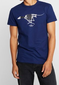 Námorně modré bavlněné tričko s grafickým potiskem kostry dinosaura oblečeného v černém oděvu, s bílým textem níže. Krátké rukávy, kulatý výstřih.