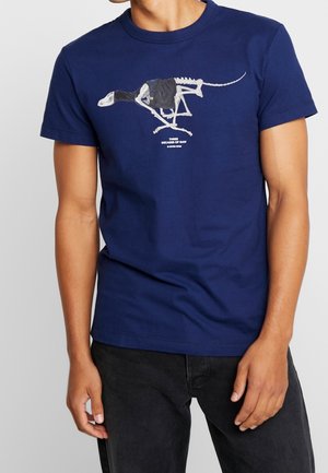 Camiseta de algodón azul marino con un diseño gráfico de un esqueleto de dinosaurio vestido con una prenda negra, con texto blanco debajo. Mangas cortas, cuello redondo.