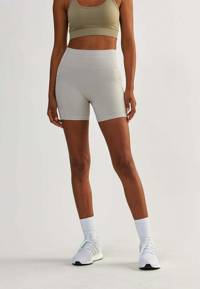 Soutien-gorge de sport beige et short de vélo taille haute assorti, fabriqué dans un tissu lisse et extensible. Des chaussures de sport blanches et des chaussettes complètent le look.