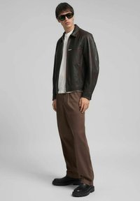 Giacca di pelle marrone con chiusura a zip, abbinata a una maglietta bianca e pantaloni marroni. Occhiali da sole neri e scarpe nere robuste completano il look.