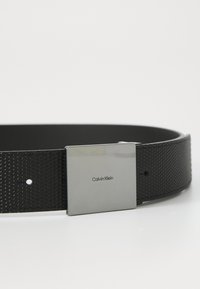 Zwarte textuurriem met een zilveren rechthoekige gesp gegraveerd met "Calvin Klein", met een gladde binnenkant en verstelbare gaten.