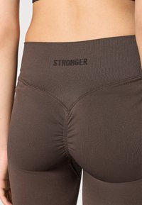 Leggings marroni a vita alta con texture a coste e dettagli arricciati sul retro. La cintura presenta la parola "STRONGER" in nero.