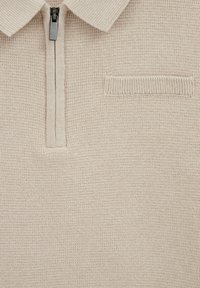 Polo en maille beige avec col à fermeture éclair partielle, tissu texturé et petit détail de poche horizontale sur la poitrine.