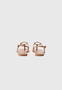Sandali piatti marroni con sottili cinghie incrociate, design a strappo per le dita e una texture in pelle liscia. Presentano dettagli metallici nella parte anteriore.