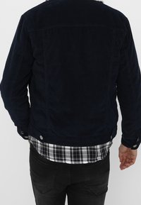 Marinblå kordflortjaket med knäppte detaljer, med en klassisk krage och en rutig skjorta synlig under.