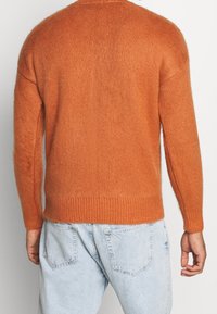 Pomarańczowy dziany sweter z długimi rękawami, prążkowanym dołem oraz luźnym krojem. Miękka tekstura i puszysta powierzchnia, noszony na jasnoniebieskich dżinsach.