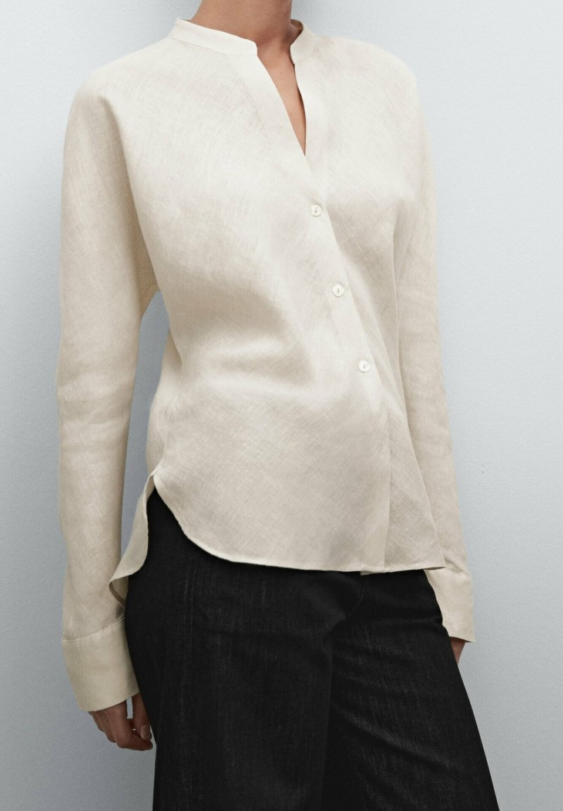Femme portant un chemisier crème boutonné à manches longues, légèrement rentré dans un pantalon noir, sur un fond uni.
