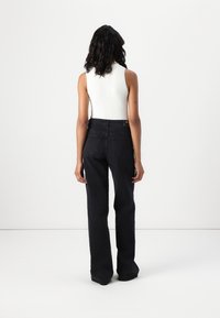 Femme debout de dos, portant un haut blanc sans manches à côtes et un jean noir taille haute, sur un fond uni.