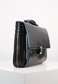 Borsa in pelle nera con cuciture blu a contrasto, dotata di chiusura a patta, hardware in argento e due cerniere laterali per ulteriore spazio di archiviazione.
