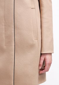 Beige jas met een gladde textuur, voorzien van een zwarte verticale naad, zijzakken en een getailleerde pasvorm, die de minimalistische ontwerpelementen benadrukt.