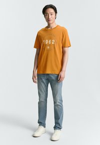 Camiseta naranja de manga corta con gráfico circular que presenta "1962" en gris claro, combinada con jeans azul claro y zapatillas blancas.