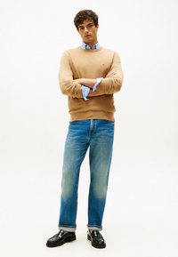 Pulover tricotat bej, cămașă albastră deschis cu guler dedesubt, blugi aliniați relaxați de culoare albastră și pantofi loafers negri. Un ansamblu simplu și clasic.