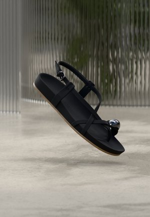 Sort sandal med krydsede stropper, flad sål og en dekorativ metalaccent. Glat mat tekstur og lys brun ydersål.