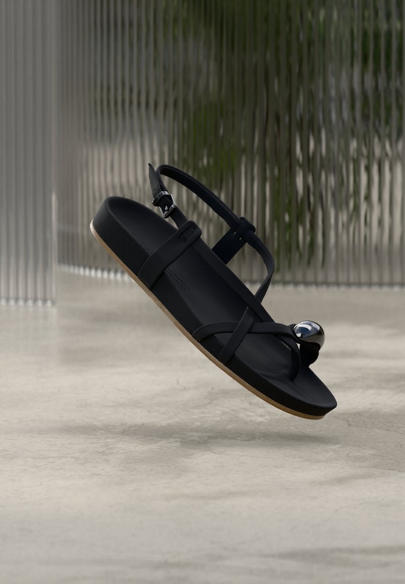 Sandale neagră cu barete încrucișate, talpă plată și un accent metalic decorativ. Textură mată netedă și talpă exterioară de culoare maro deschis.