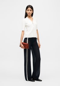 Γυναίκα που φοράει λευκό τοπ με κοντά μανίκια, μπλε σκούρο παντελόνι με φαρδιά μπατζάκια και λευκές ρίγες, κρατώντας κόκκινο τσαντάκι, στέκεται μπροστά σε απλό φόντο.