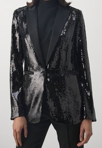 Blazer negro y plateado adornado con lentejuelas, con solapa de satén, que presenta dos bolsillos frontales y un diseño ajustado, texturizado con detalles brillantes de lentejuelas.