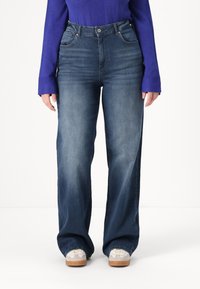 Weite Denim-Jeans in mittlerer Waschung; ausgestattet mit fünf Taschen, Gürtelschlaufen und einem Knopfverschluss. Getragen mit grauen und cremigen Sneakers.