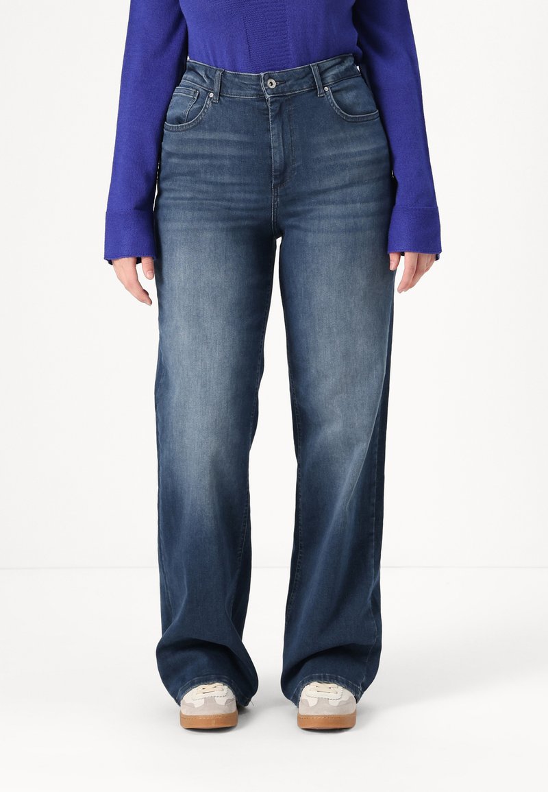 Breda jeans i medium tvätt; har fem fickor, bältesöglor och knäppning med knappar. Bärs med grå och krämfärgade sneakers.