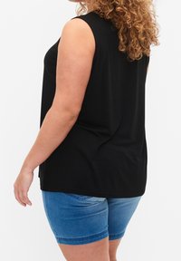 Osoba z kręconymi włosami, ubrana w czarną bluzkę bez rękawów i niebieskie szorty dżinsowe, sfotografowana z tyłu na tle białego tła.