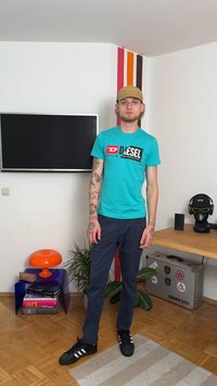 Τυρκουάζ μπλουζάκι με λογότυπο "DIESEL", ναυτικές παντελόνες, μαύρα αθλητικά παπούτσια. Διαθέτει τατουάζ στα χέρια, στέκεται σε δωμάτιο με πολύχρωμες επιφάνειες.