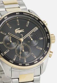 Montre chronographe avec un cadran noir, des accents dorés, un boîtier et un bracelet en acier inoxydable, une échelle tachymétrique, trois sous-cadrans et un logo crocodile.