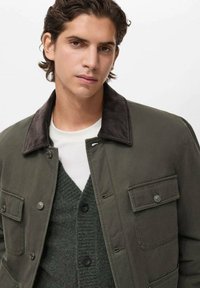 Giovane uomo con capelli scuri di lunghezza media che indossa una giacca verde oliva con tasche abbottonate e un cardigan lavorato a maglia verde scuro sopra una camicia bianca.