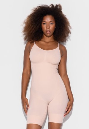 Vrouw met volumineus krullend haar, gekleed in een nauwsluitend, mouwloos bodysuit in naaktkleur, staand tegen een eenvoudige lichte achtergrond.