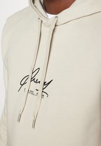 Sudadera beige hecha de una suave mezcla de algodón, con capucha ajustable y un logotipo negro bordado en el pecho. Textura suave con terminales de metal.