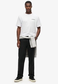 Hvid bomulds t-shirt med et lille sort "SUPERDRY" logo, sorte bukser og en grå sweatshirt bundet om livet; sorte sneakers med beige såler.
