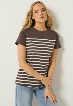 Blonde Frau trägt ein dunkelbraun-weiß gestreiftes kurzärmeliges Oberteil mit Knopfdetails an den Schultern und hellblaue Jeans.