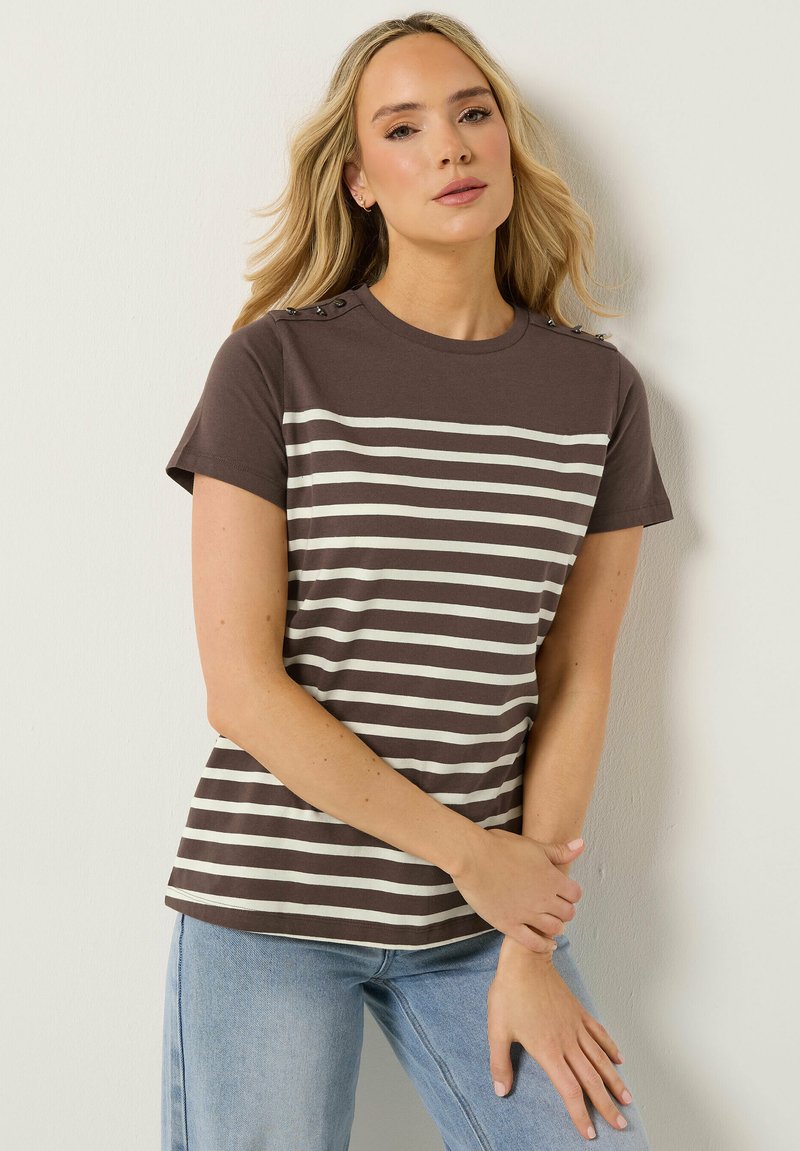 Femme blonde portant un haut à manches courtes rayé marron foncé et blanc avec des détails de boutons sur les épaules et un jean en denim bleu clair.