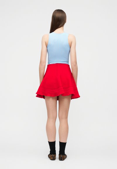 Jeune femme aux longs cheveux bruns portant un haut sans manches à rayures bleues et blanches, une jupe plissée rouge, des chaussettes noires et des ballerines imprimé léopard, debout.