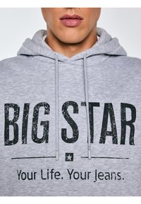 Inimese lähivaade, kellel on seljas helehall kapuutsiga pusa, mille esiküljel musta tekstiga "BIG STAR" ja "Your Life. Your Jeans.".