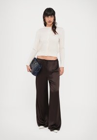 Γυναίκα που φοράει λευκό κοντό ζακετάκι, φαρδιά μαύρη σατέν παντελόνα, λευκά μυτερά παπούτσια, κρατώντας μαύρη τσάντα χειρός μπροστά σε απλό φόντο.