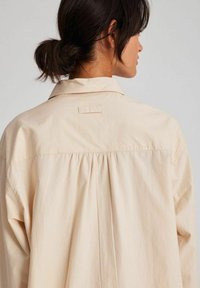 Camicia beige chiaro con colletto classico e dettaglio plissettato nella parte posteriore, realizzata in tessuto morbido. Maniche lunghe con polsini a bottone, che mostrano un design pulito.