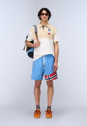 Ung mand iført solbriller, beige og hvid polo med mærker, blå shorts med flagdesign, orange sneakers og bærer en rygsæk på den ene skulder.