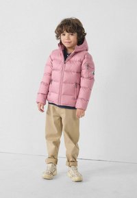 Chaqueta tipo plumas de color rosa claro con capucha, cierre frontal con cremallera y dos bolsillos laterales. Combinada con pantalones caqui y zapatillas beiges. Diseño simple y limpio.